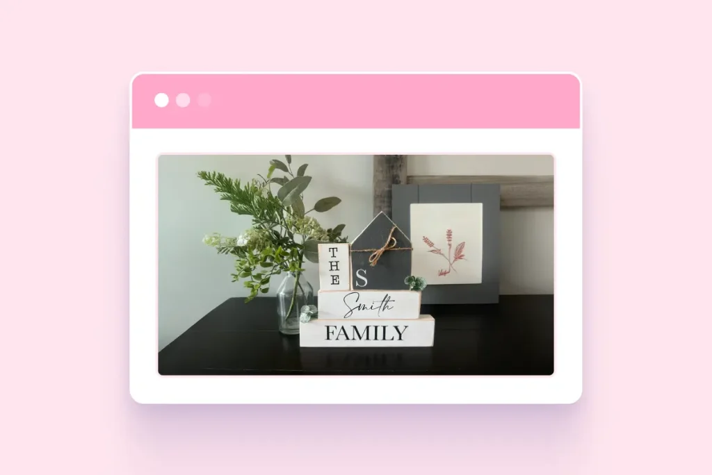 A personalized wooden home decor set on a dark tabletop, featuring stacked white and grey blocks that read "THE Smith FAMILY" alongside a small house-shaped block, a glass vase with greenery, and a framed botanical print