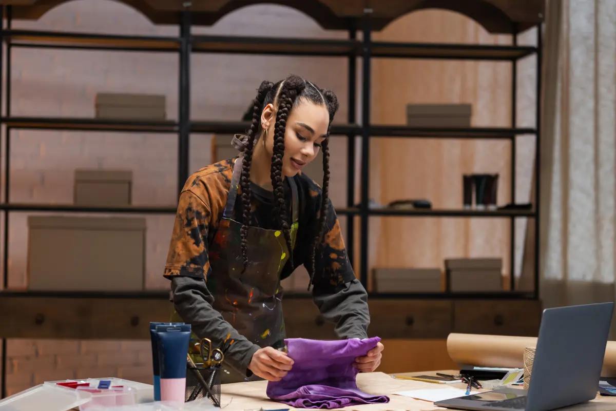Woman in a paint-splattered apron folding a purple garment at a workbench with craft supplies and a laptop nearby
