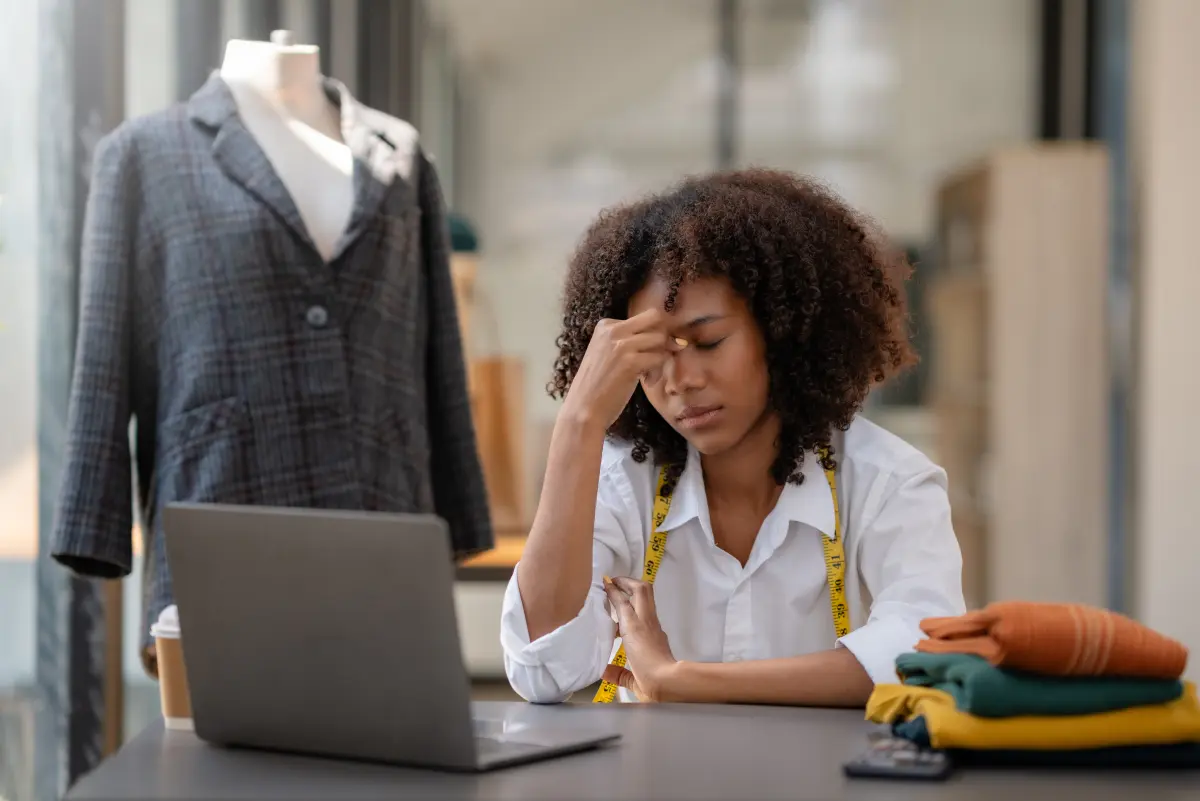 Stressed designer sitting at a desk with her head in her hand, surrounded by a laptop, folded fabrics, and a tailored jacket on a mannequin
