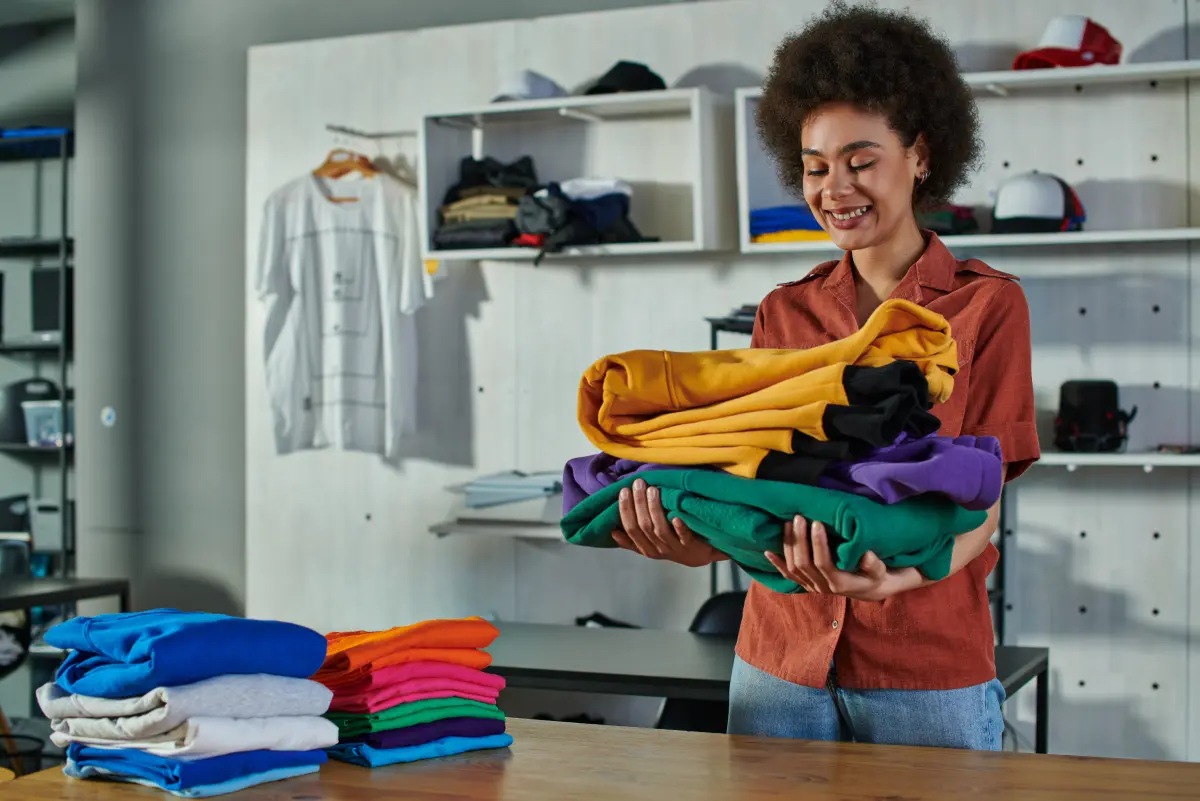 Smiling woman holding a stack of colorful folded apparel in a clothing shop with shelves of garments and caps in the background