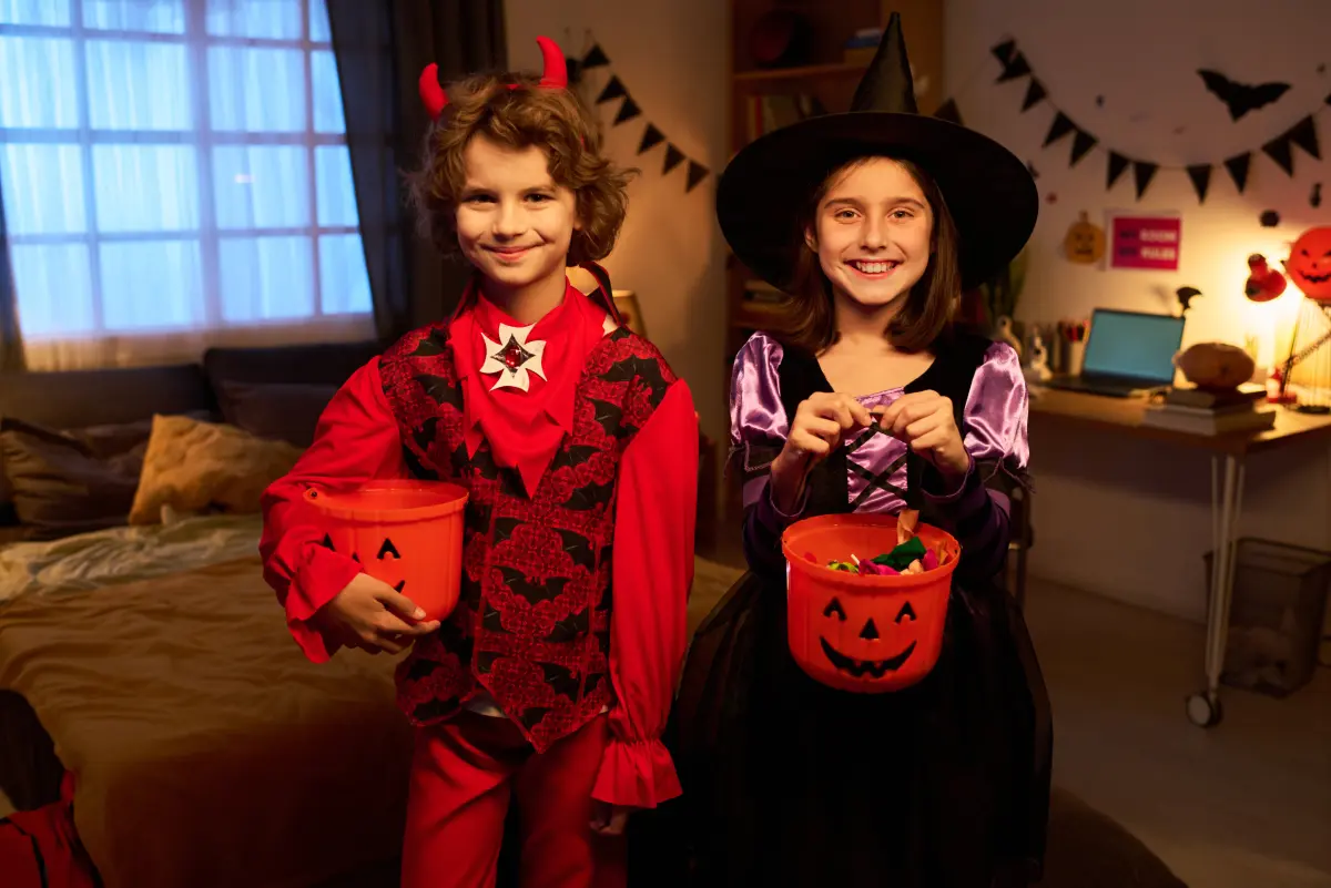 Two children in Halloween costumes, a devil and a witch, each holding a jack o lantern candy bucket in a bedroom decorated with bat and pennant garlands