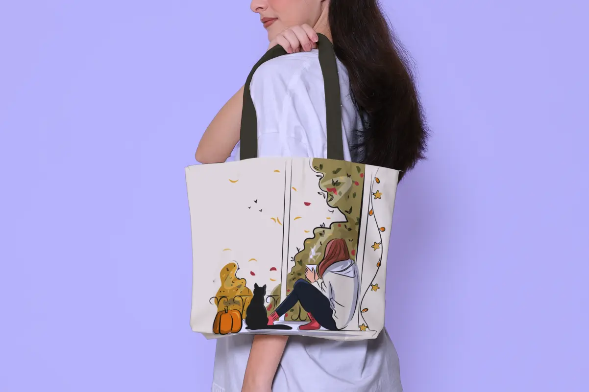 Person carrying a cream tote bag with olive straps, printed with a cozy autumn illustration of a woman reading by a window with a black cat and pumpkins.