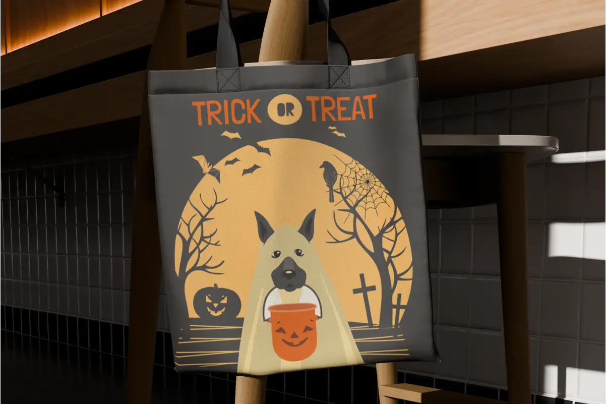Dark gray tote bag hanging on a chair, printed with "Trick or Treat" text and a dog holding a jack-o'-lantern candy pail, with bats, crosses, and a full moon graveyard scene