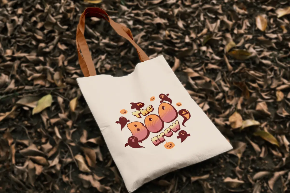 Cream canvas tote bag with orange straps, printed with retro "The Boo Crew" text surrounded by cartoon ghosts and jack-o'-lantern faces, resting on fallen autumn leaves