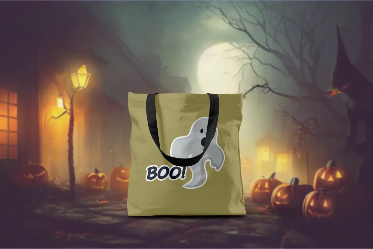 Olive-green tote bag with a minimalist ghost sticker and "Boo!" text, set against a spooky moonlit street scene with glowing jack-o'-lanterns and a silhouetted witch