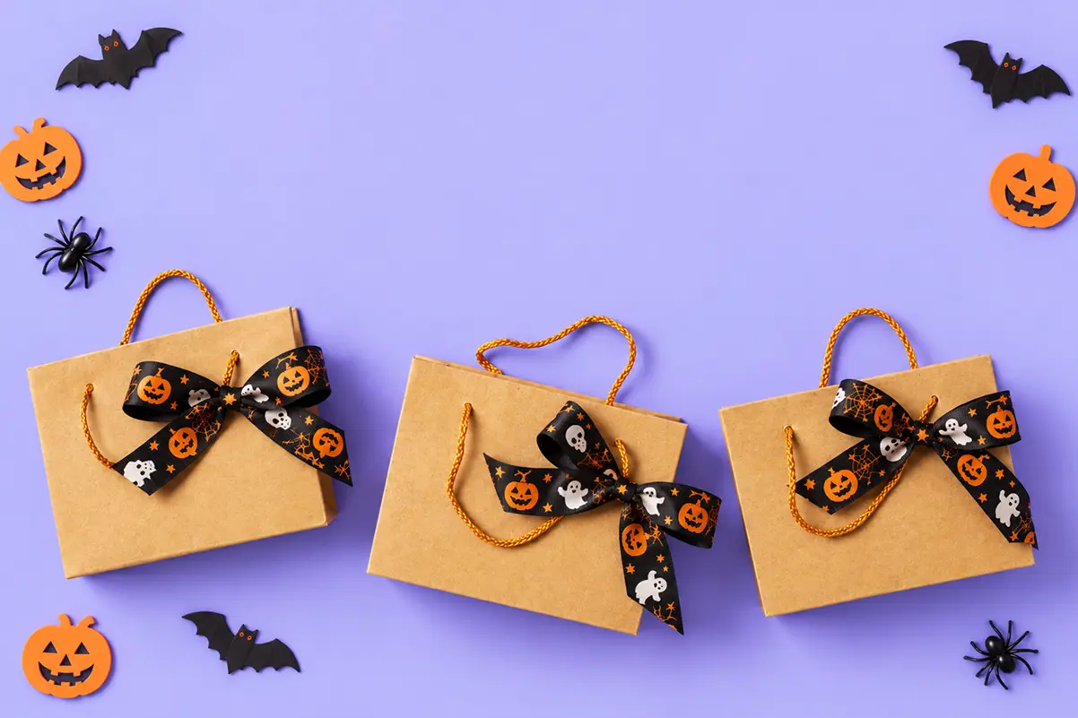 Three kraft paper gift bags tied with black-and-orange pumpkin and ghost ribbon bows, surrounded by paper pumpkin, bat, and spider decorations on a purple background