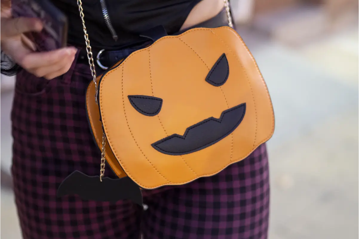 Person wearing a plaid skirt with an orange jack-o'-lantern-shaped crossbody bag featuring a carved black smiling face and a small black bat charm on a gold chain strap