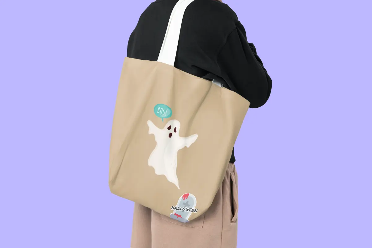 Person carrying a tan tote bag on their shoulder printed with a cartoon ghost saying "Boo!" and a bloody tombstone reading "Halloween."